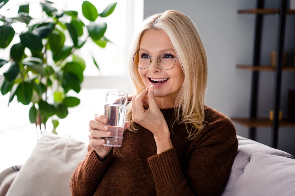 Eine lächelnde Frau mit Brille nimmt eine Tablette und hält ein Glas Wasser